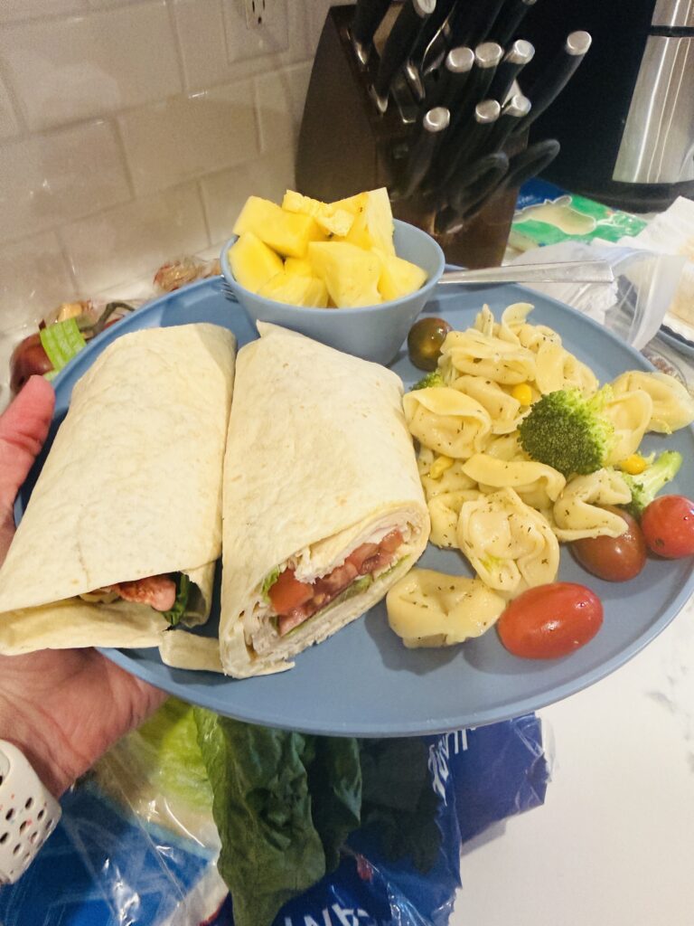 Image of the chicken club wrap sandwich on a blue plate with a small bowl of cut pineapple, and a side dish of tortellini salad with tomatoes.